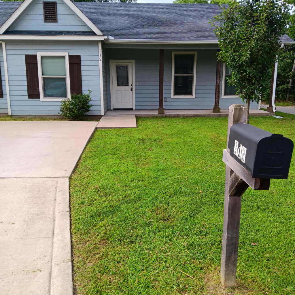 A well-maintained green lawn in front of a blue house with a black mailbox marked "112." The grass is freshly cut, and the landscaping is neat.